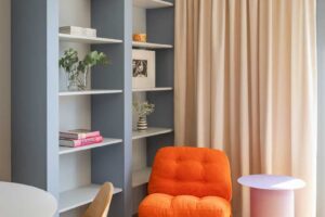Oranžové křeslo v čtecím koutě kontrastuje s modrošedou knihovnou a jemnými béžovými závěsy u okna.