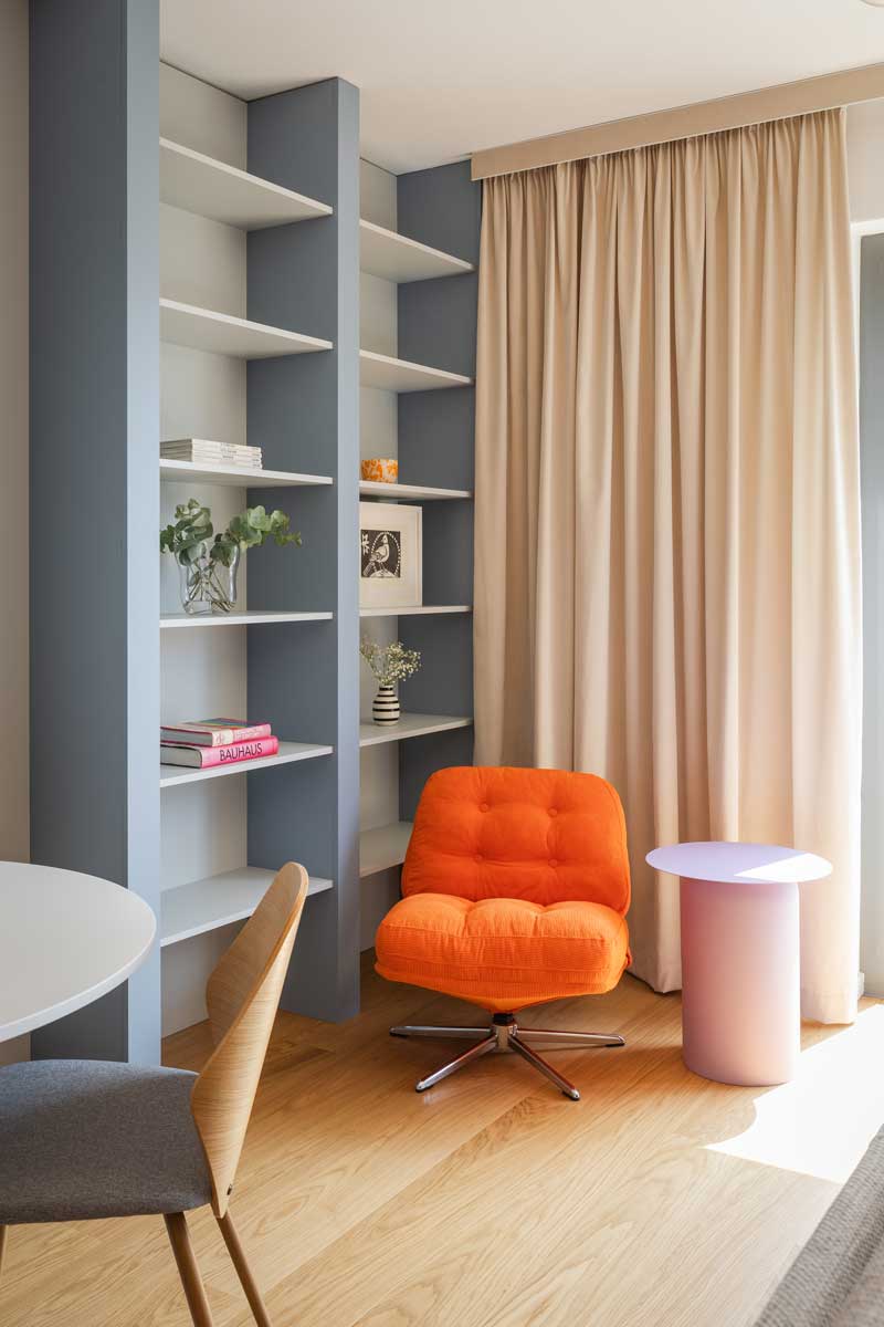 Oranžové křeslo v čtecím koutě kontrastuje s modrošedou knihovnou a jemnými béžovými závěsy u okna.