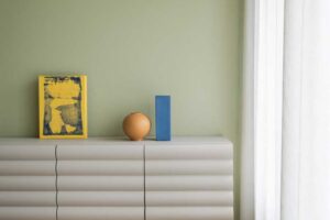 Detail komody v ložnici vyniká jemně strukturovaným nábytkem, pastelovou zelenou stěnou a dekoracemi v modré, žluté a terakotové barvě.