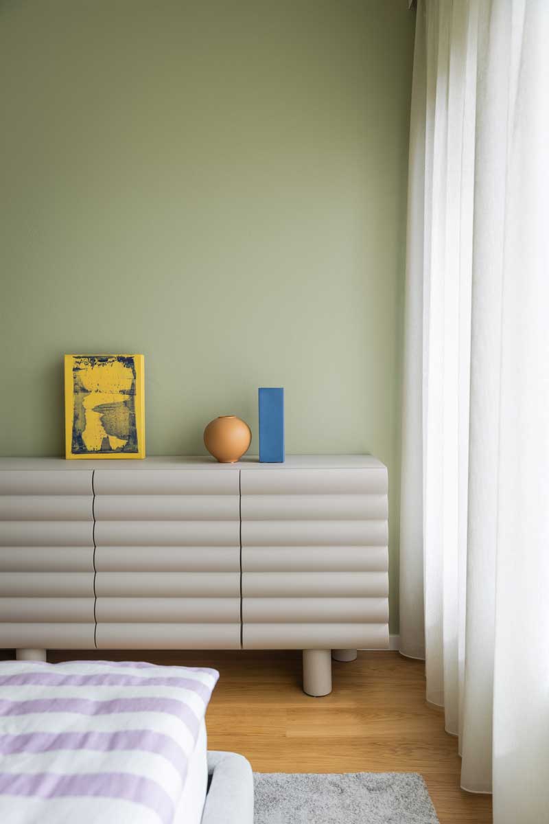 Detail komody v ložnici vyniká jemně strukturovaným nábytkem, pastelovou zelenou stěnou a dekoracemi v modré, žluté a terakotové barvě.