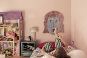 Dětský pokoj s růžovou stěnou postelí na zemi a hračkami včetně domečku pro panenky.