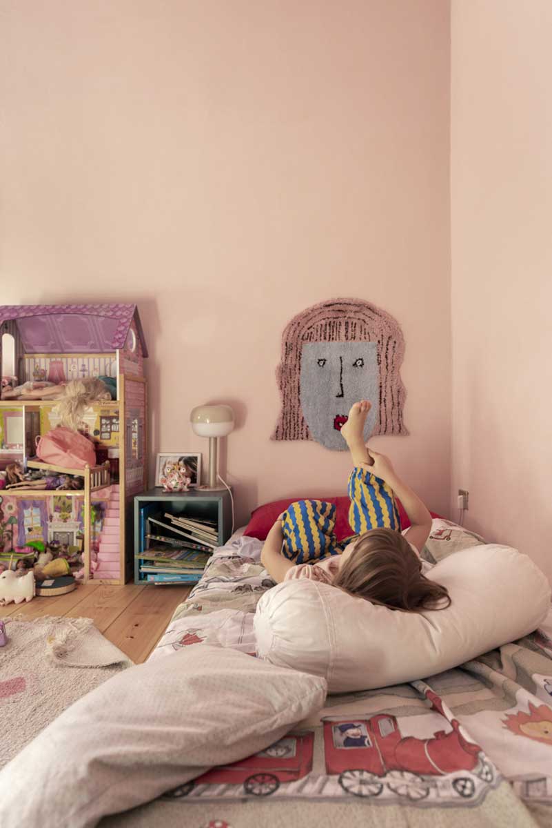 Dětský pokoj s růžovou stěnou postelí na zemi a hračkami včetně domečku pro panenky.