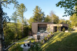 Moderní dům ve strmém svahu u jezera obklopený lesem s terasou a výhledem do krajiny.