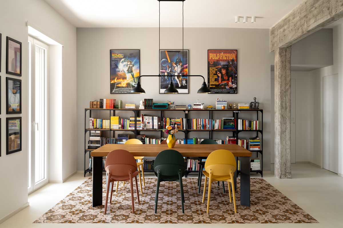 Jídelní kout s dřevěným stolem barevnými židlemi a policemi plnými knih doplněný filmovými plakáty na stěně.