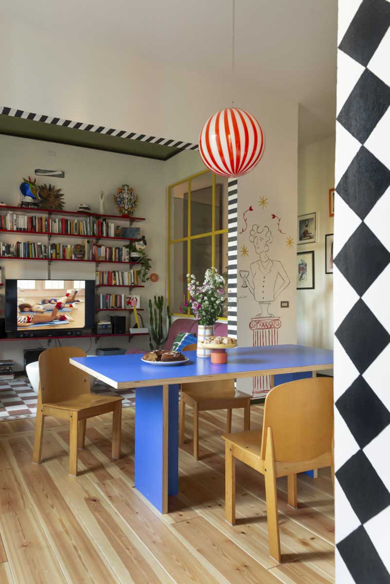 Jídelna s modrým stolem a červenobílým závěsným svítidlem propojená s obývacím pokojem plným knih.