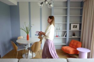 Jídelní kout s kulatým stolem a dřevěnými židlemi navazuje na vestavěnou knihovnu a modrošedé panely, které propojují kuchyň a obývací část.