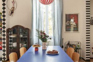 Modrý jídelní stůl se světlými dřevěnými židlemi pod výrazným červenobílým závěsným svítidlem.