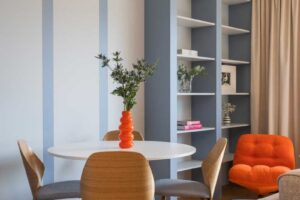 Kulatý jídelní stůl se čtyřmi dřevěnými židlemi stojí před modrošedými panely a otevřenou knihovnou, které dodávají interiéru rytmus.