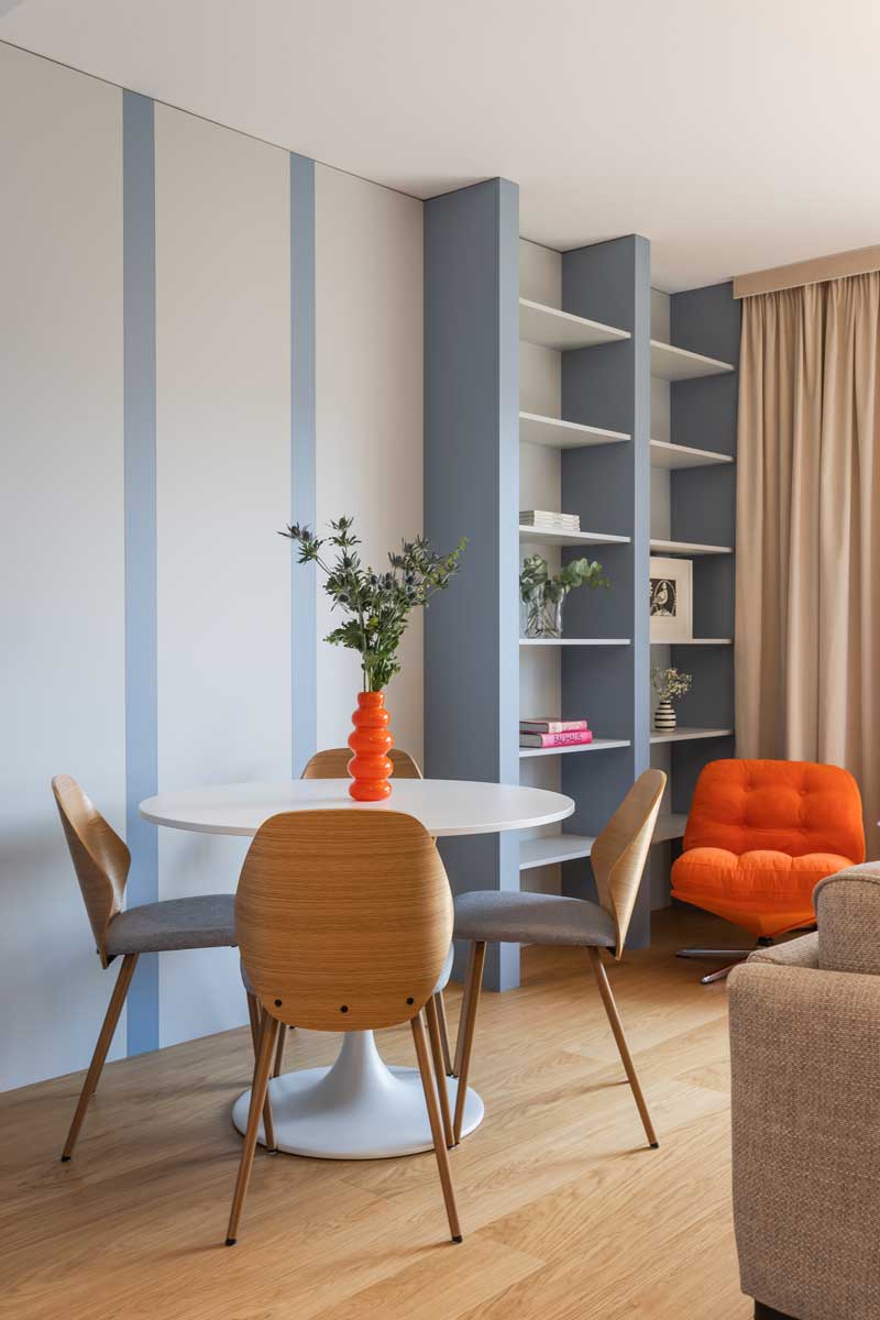 Kulatý jídelní stůl se čtyřmi dřevěnými židlemi stojí před modrošedými panely a otevřenou knihovnou, které dodávají interiéru rytmus.