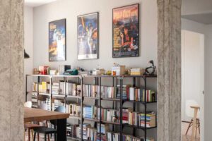Otevřená knihovna za jídelním stolem která propojuje úložný prostor pro knihy s dekoracemi a filmovými plakáty na stěně.