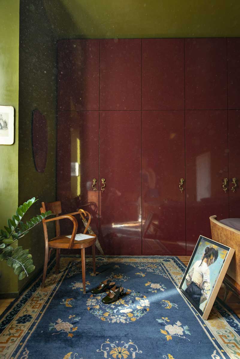 Ložnice s olivově zelenou stěnou bordó skříní a vintage nábytkem doplněná modrým orientálním kobercem.