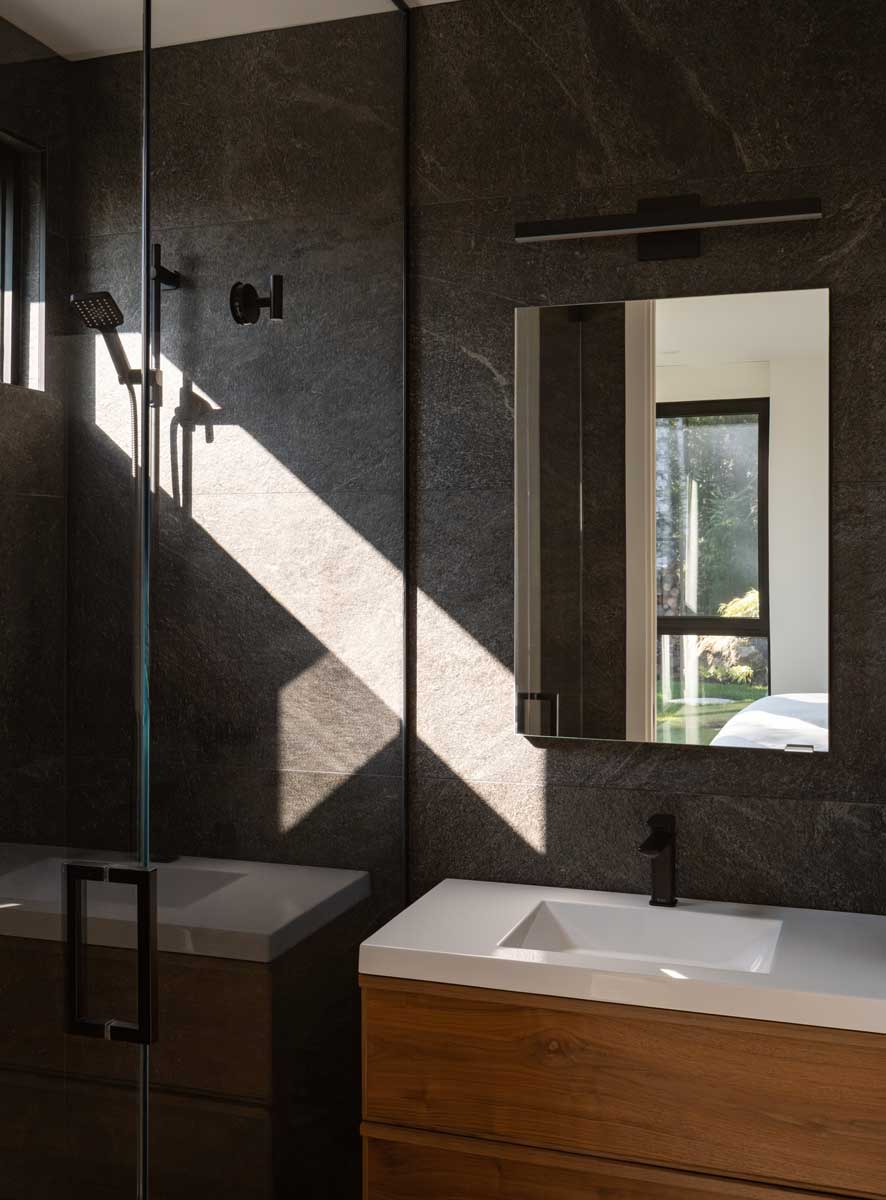 Moderní koupelna s tmavým obkladem, sprchovým koutem a minimalistickým umyvadlem.