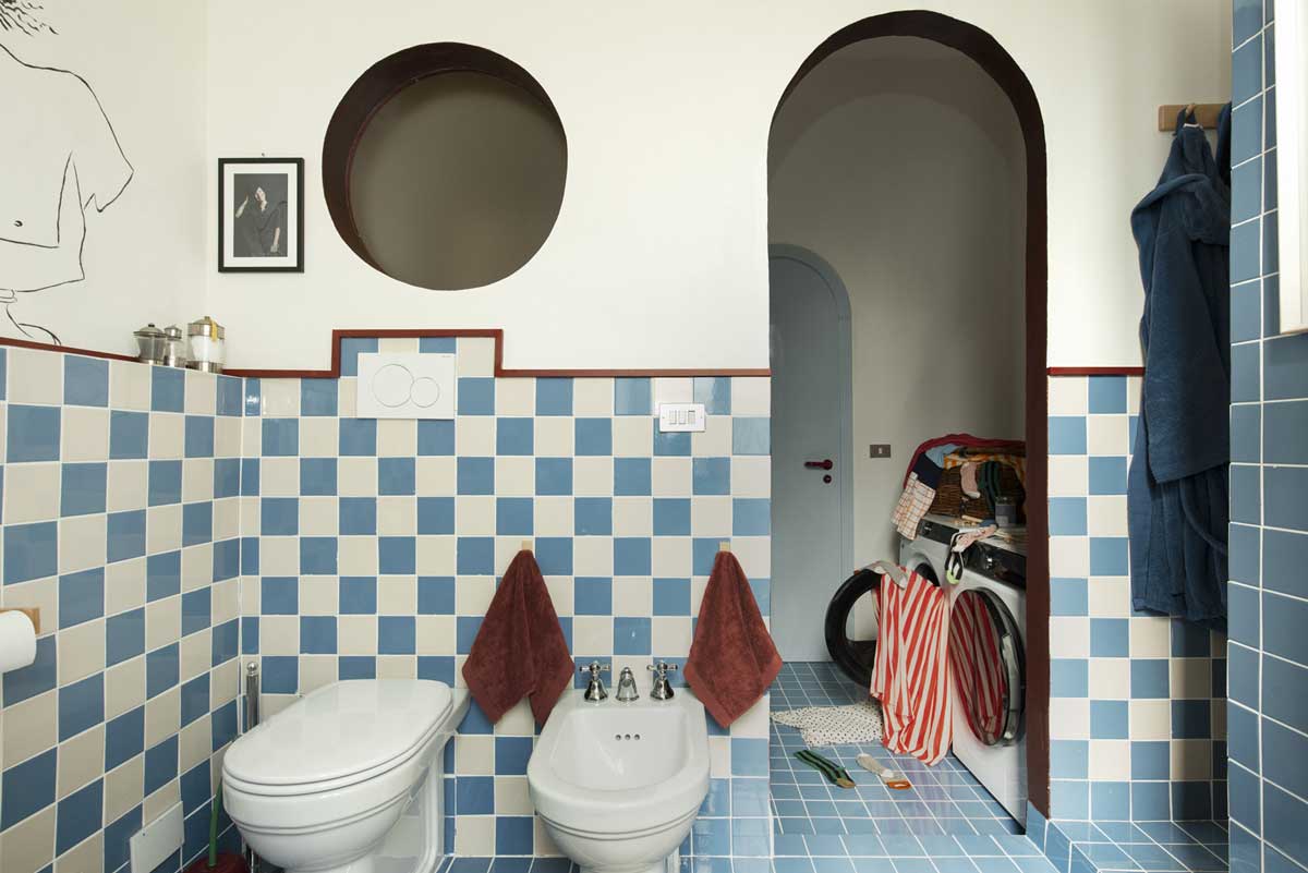 Koupelna s modrobílými obklady a obloukovým průchodem vedoucím do malé prádelny s pračkou.
