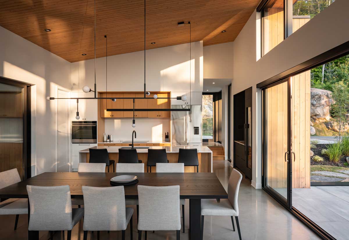 Otevřený interiér s jídelnou, kuchyní a vysokým stropem s velkými okny.