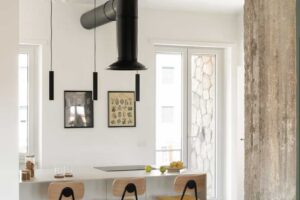 Minimalistická kuchyně s ostrůvkem doplněná ručně vyráběnou kovovou digestoří a subtilními závěsnými svítidly nad pracovní plochou.