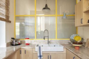 Světlá kuchyň s dřevěnými skříňkami a žlutými rámy prosklené příčky nad dřezem.