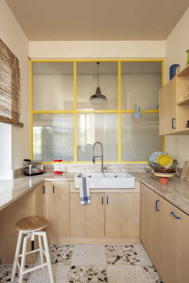 Světlá kuchyň s dřevěnými skříňkami a žlutými rámy prosklené příčky nad dřezem.