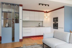 Bílá kuchyňská linka s vestavěnými spotřebiči je doplněna barevným soklem a kontrastní podlahou.