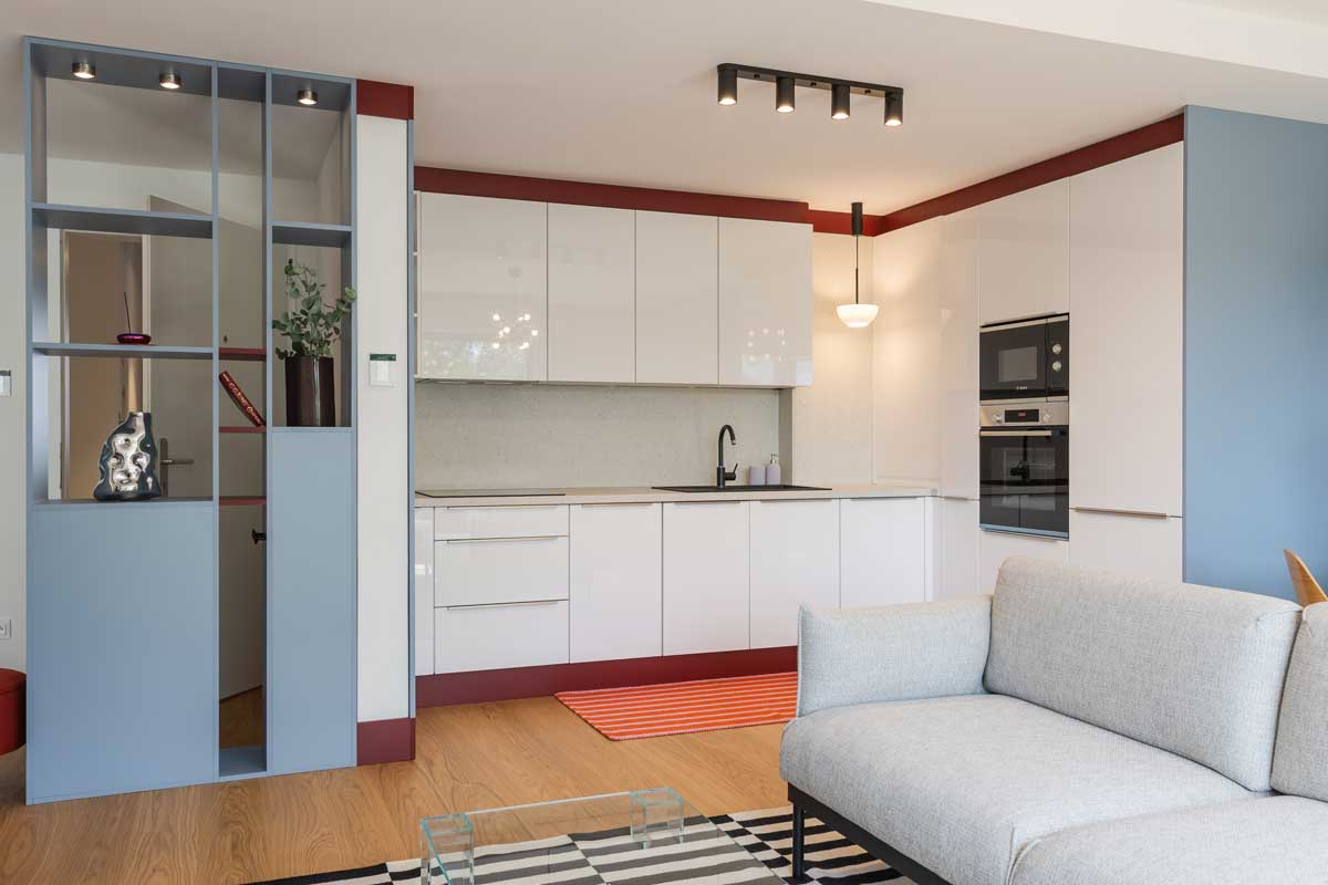 Bílá kuchyňská linka s vestavěnými spotřebiči je doplněna barevným soklem a kontrastní podlahou.