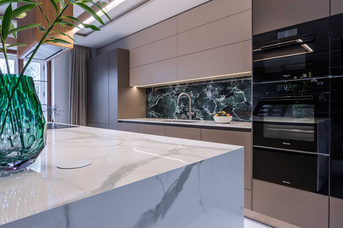 Kuchyňský ostrůvek obložený keramikou s dekorem kamene doplněný světlou pracovní deskou a designovým osvětlením.