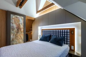 Ložnice v podkroví s vestavěnými úložnými prostory a čalouněným čelem postele v kombinaci dřeva a neutrálních tónů.