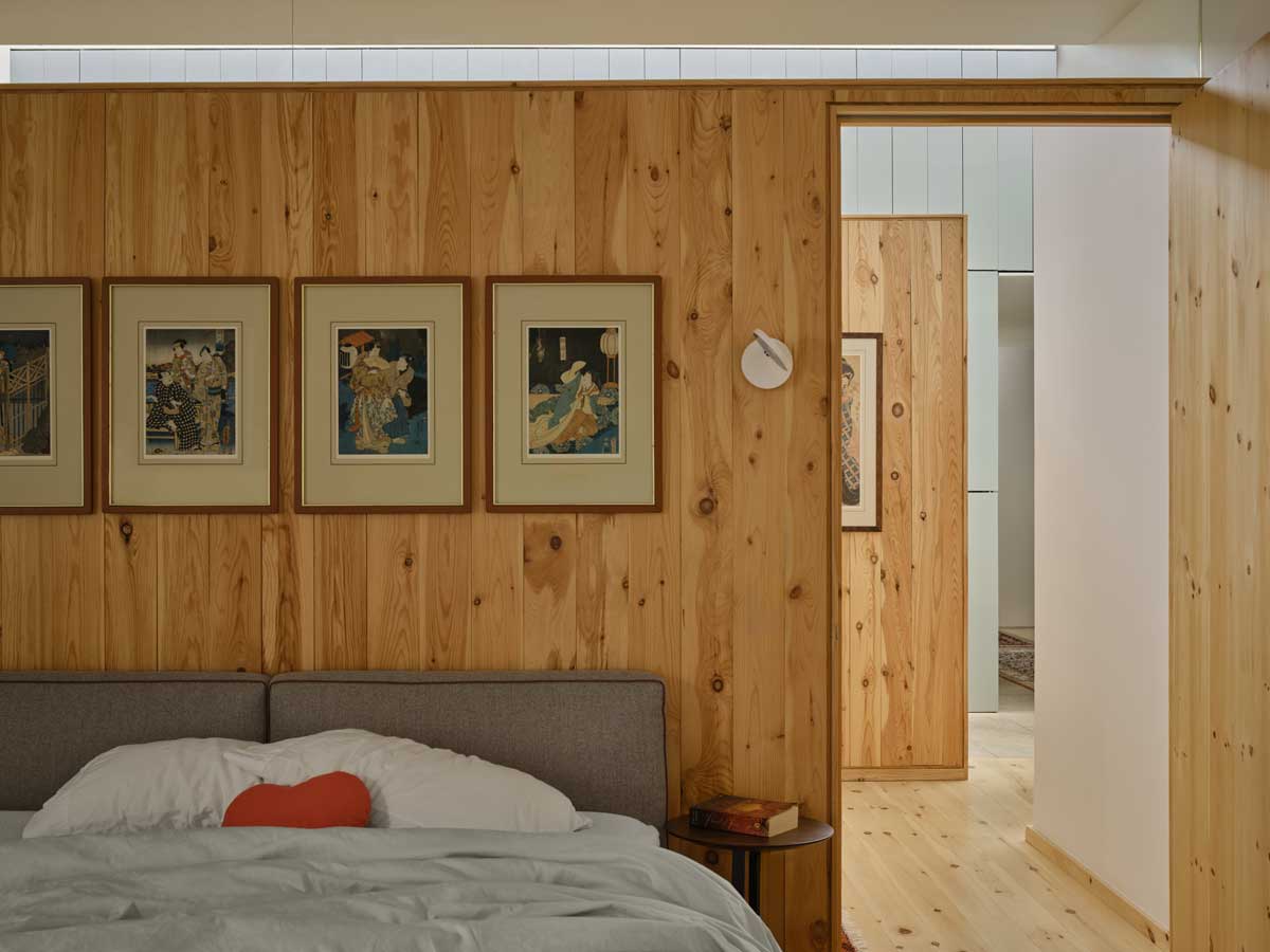 Ložnice s dřevěným obkladem, jednoduchou postelí a galerií obrazů nad čelem