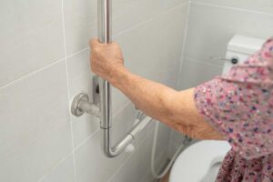 Senior se opírá o bezpečnostní madlo u toalety které pomáhá při vstávání a pohybu v koupelně.