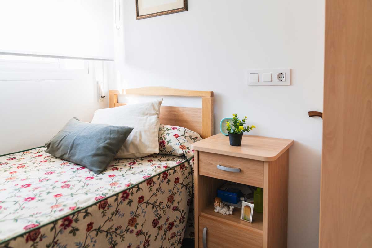 Nízká postel a noční stolek v ložnici které umožňují seniorovi snadné vstávání a bezpečný pohyb.