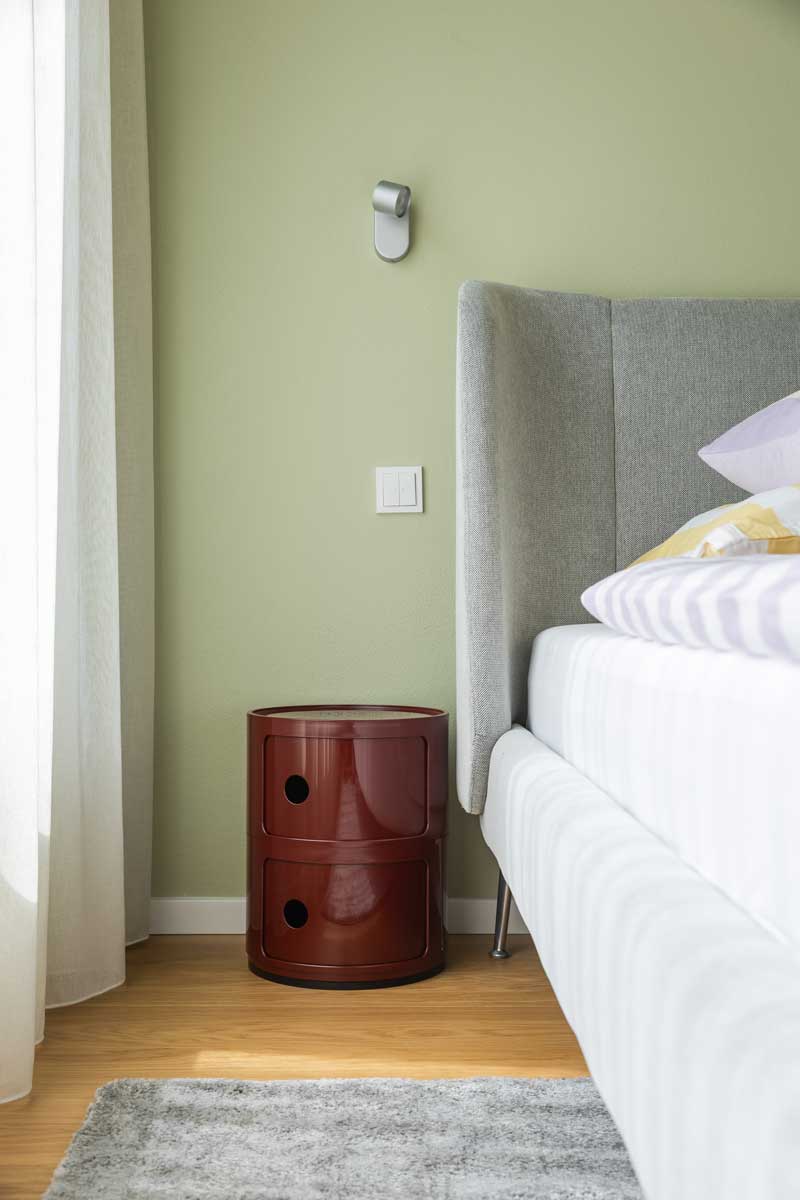 Noční stolek v sytě červeném odstínu stojí vedle čalouněné postele a kontrastuje s jemně zelenou stěnou ložnice.