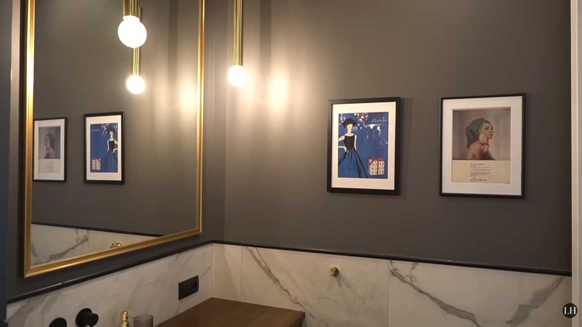 Detail koupelny s velkým zrcadlem, nástěnnými světly a dekorativními obrazy na tmavé stěně.