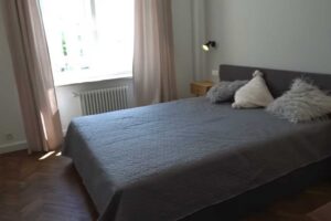 Ložnice po rekonstrukci s velkou postelí, světlými závěsy a renovovanými parketami.