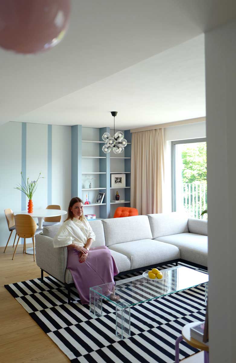 Designérka sedí v obývacím prostoru, který propojuje jídelní kout, knihovnu a světlou sedací soupravu.