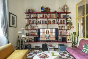 Obývací pokoj s policovou knihovnou plnou knih retro nábytkem a barevnými doplňky.