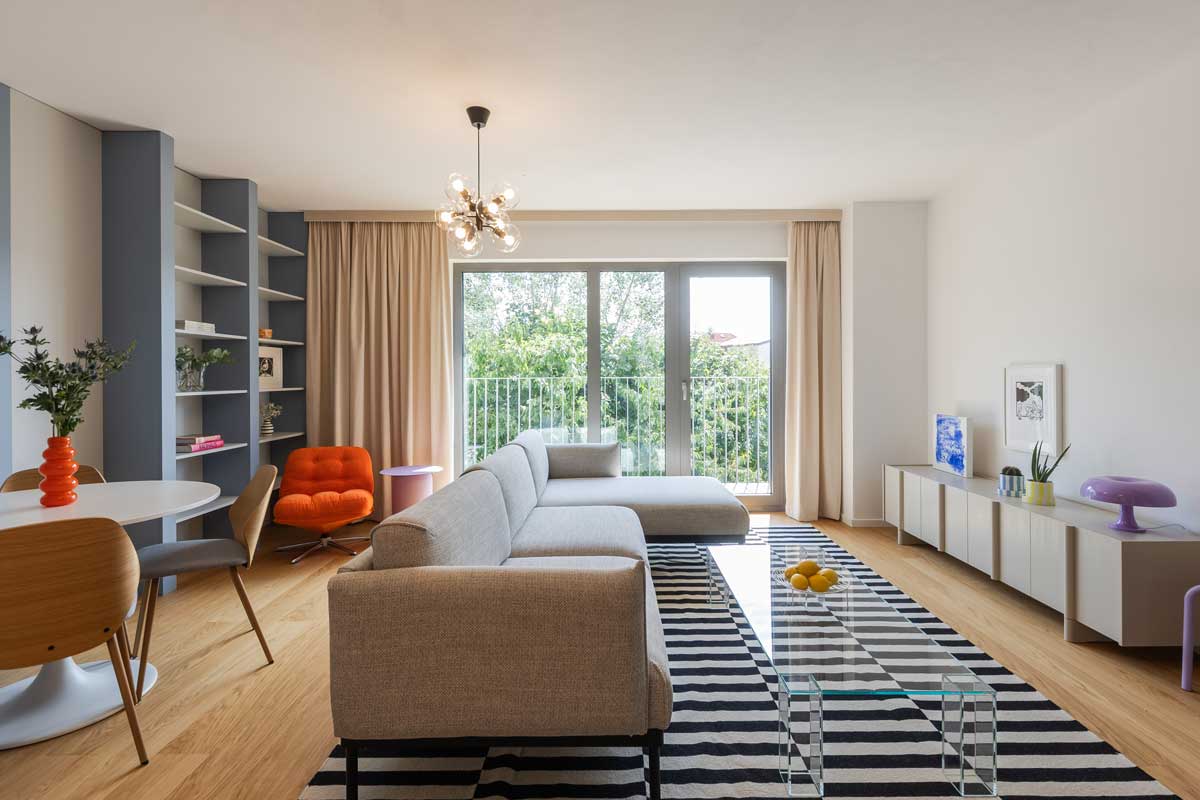 Světlý obývací pokoj s velkým oknem a balkonem je doplněn výrazným kobercem, skleněným stolkem a oranžovým křeslem.