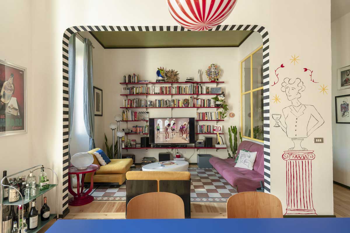 Pohled skrz obloukový průchod do obývacího pokoje s knihovnou růžovou pohovkou a výraznou nástěnnou kresbou.