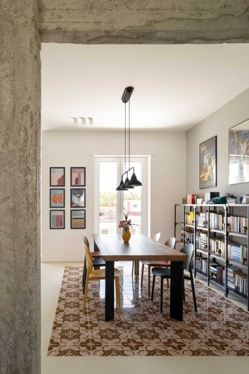 Jídelna s velkým dřevěným stolem barevnými židlemi a historickou dlažbou která vymezuje prostor v otevřeném interiéru.