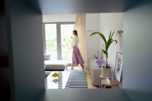 Pohled skrz vestavěnou knihovnu odhaluje světlý obývací prostor s balkonem, minimalistickou komodou a pastelovými doplňky.