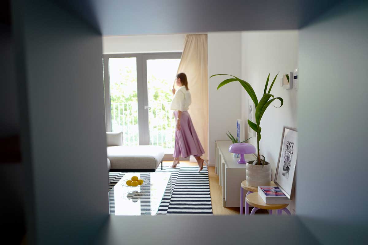 Pohled skrz vestavěnou knihovnu odhaluje světlý obývací prostor s balkonem, minimalistickou komodou a pastelovými doplňky.