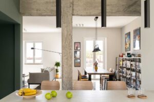 Pohled z kuchyňského ostrůvku do otevřené denní zóny kde se setkává jídelna obývací pokoj a přiznaná betonová konstrukce.