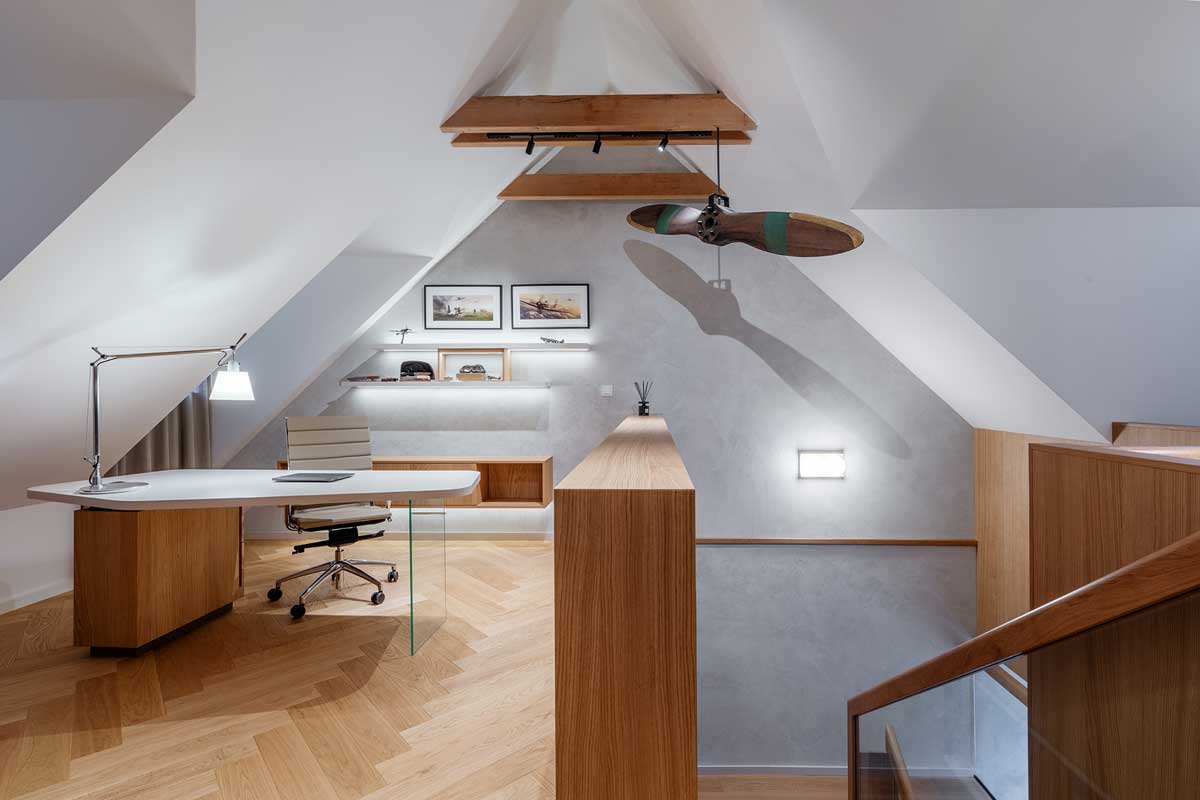 Podkrovní pracovna s masivní dřevěnou podlahou šikmými stropy a vestavěnými úložnými prostory.