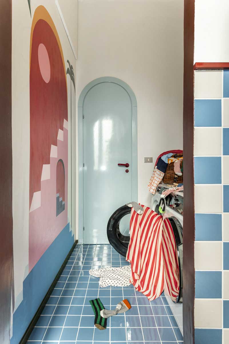 Úzká prádelna s modrou dlažbou a ručně malovanou nástěnnou dekorací na stěně.
