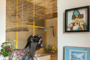 Majitelka bytu stojí u kuchyňského pultu se žlutými rámy prosklené příčky a dekoracemi na stěně.