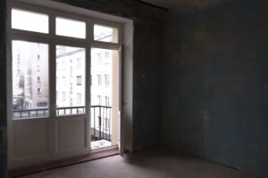 Obytný pokoj před rekonstrukcí s vysokými stropy a balkonovými dveřmi typickými pro staré činžovní domy.