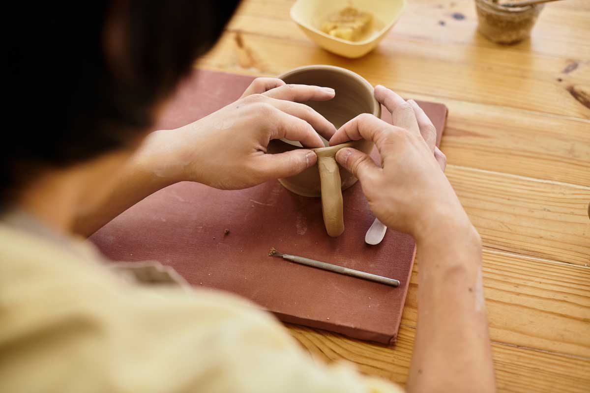 Keramik tvaruje hrnek z hlíny na pracovním stole a ručně připevňuje jeho ucho.
