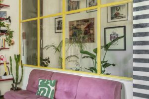 Růžová pohovka stojí před prosklenou příčkou se žlutými rámy a rostlinami v obývacím pokoji.