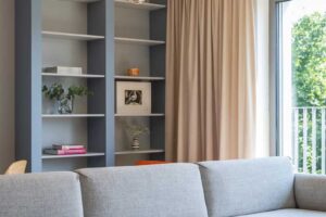 Světlá sedací souprava stojí v centru obývací části a doplňuje ji skleněný stolek, výrazný koberec a knihovna v modrošedém odstínu.