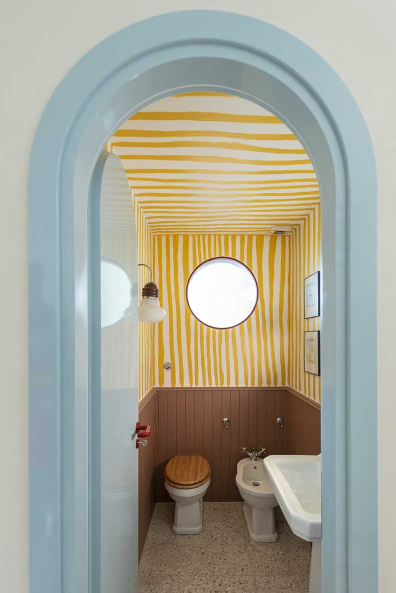Pohled skrz obloukový průchod do koupelny se žlutě pruhovanými stěnami a bílou sanitou.