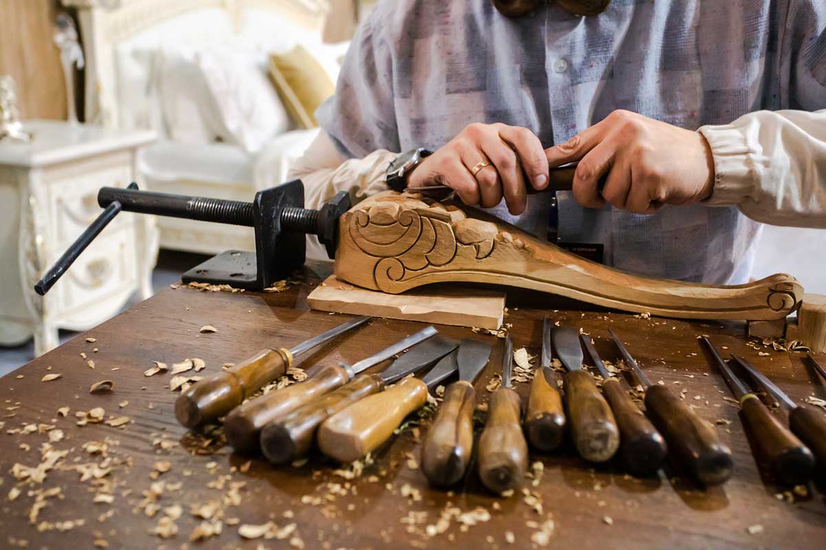 Řezbář vyřezává ozdobný detail do dřevěné nohy nábytku a používá tradiční dláta.
