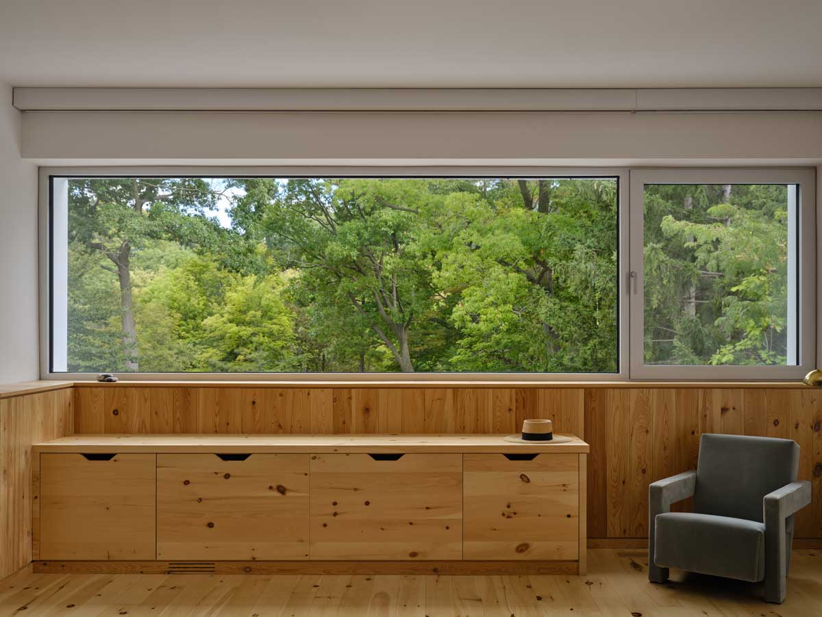 Vestavěná dřevěná skříň pod oknem s výhledem do zeleně a jednoduchým křeslem vedle