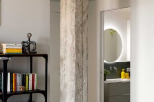 Přiznaný betonový sloup odděluje obytnou část bytu od vstupu do koupelny kde dominuje kulaté zrcadlo s podsvícením a minimalistická skříňka s umyvadlem.